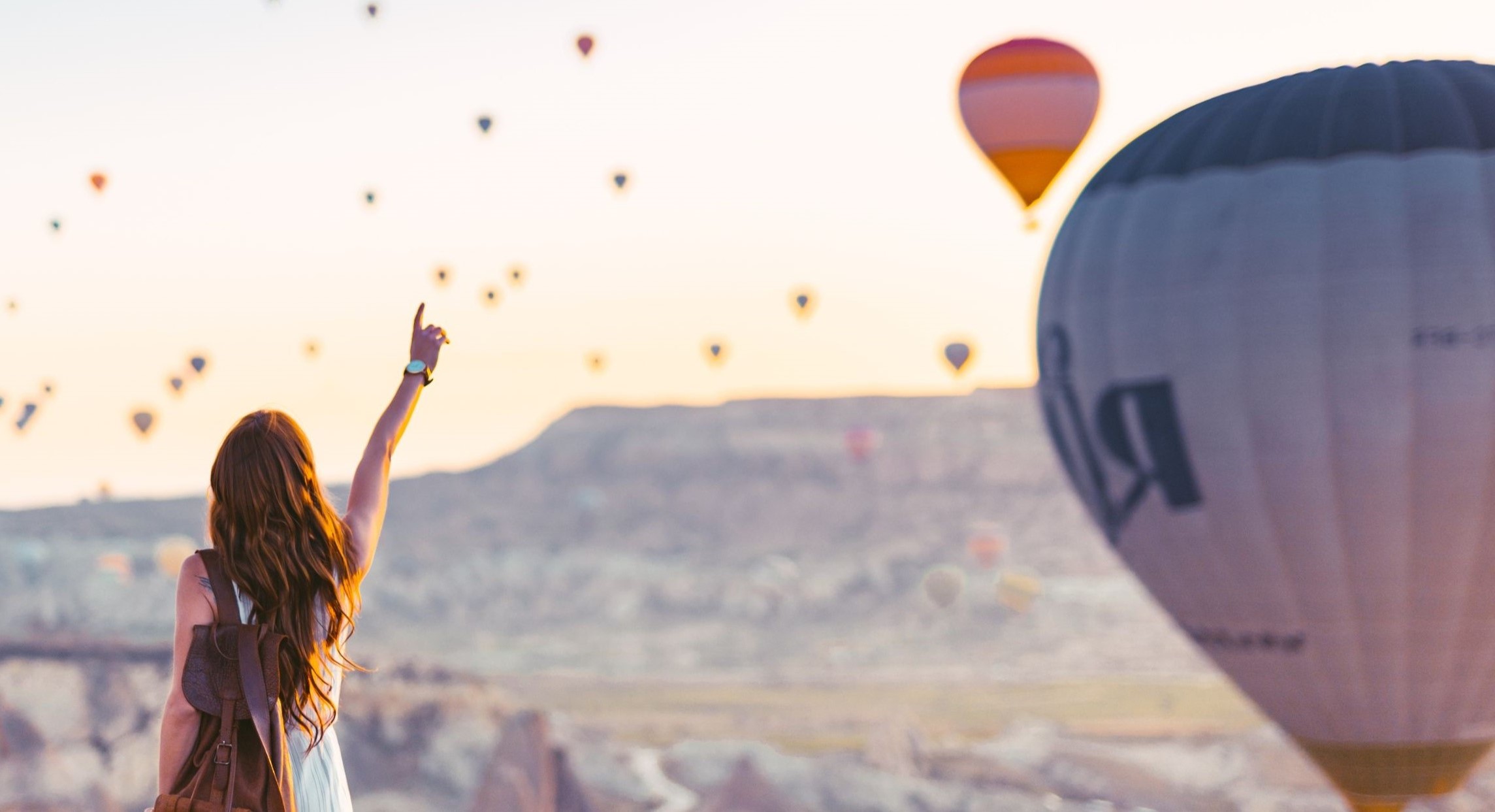 Frau mit weißem Kleid steht vor einer bergigen Landschaft. Am Himmel sind zahlreiche Heißluftballons zu sehen. Die Frau winkt ihnen mit einem Arm zu.