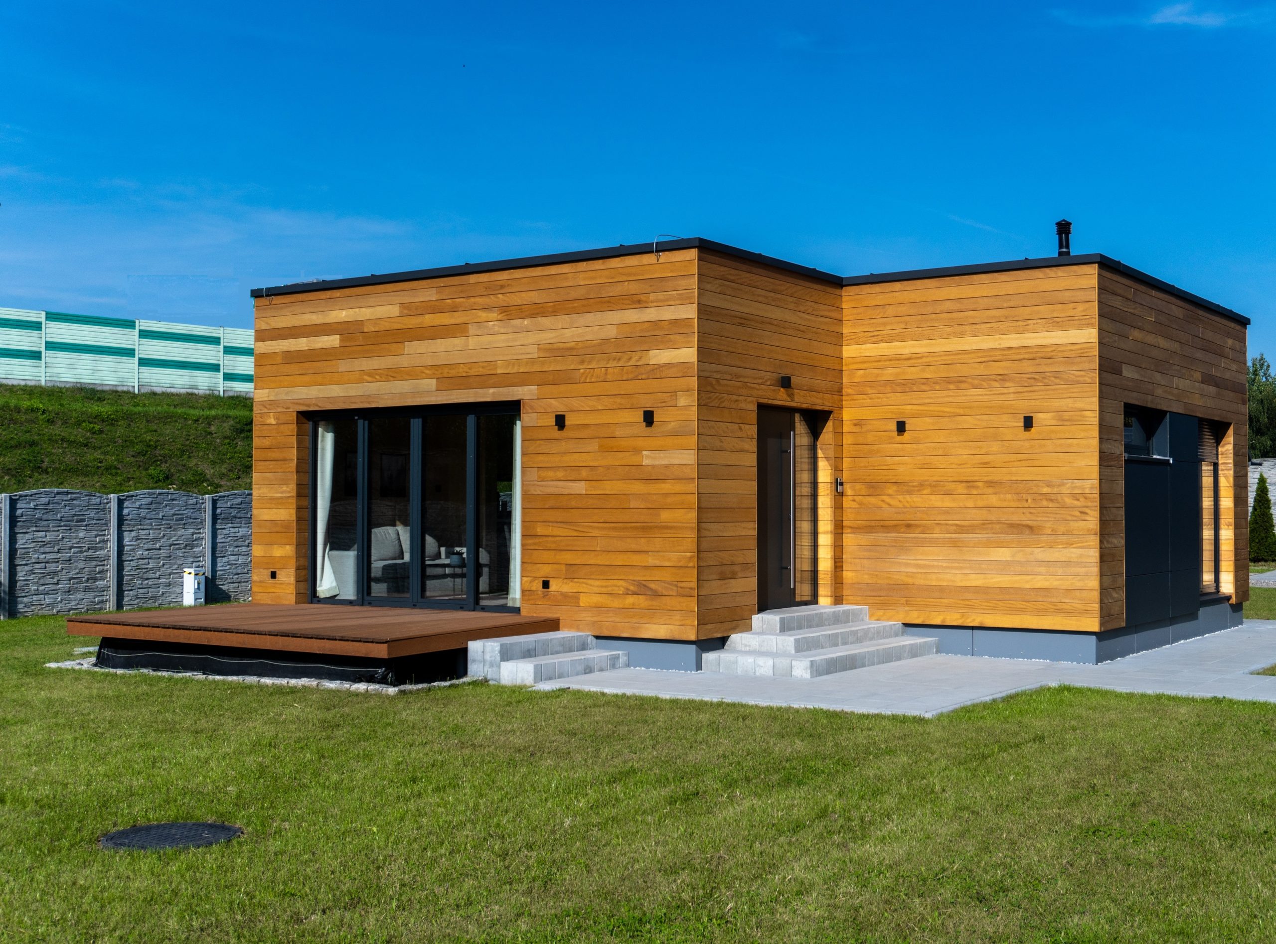 Außenansicht des Modulhauses Vang mit einer Holzfassade. Das Haus steht auf einem Grundstück mit grünem Rasen, und vor dem Haus erstreckt sich eine Holzterrasse. Das Wetter ist sonnig.