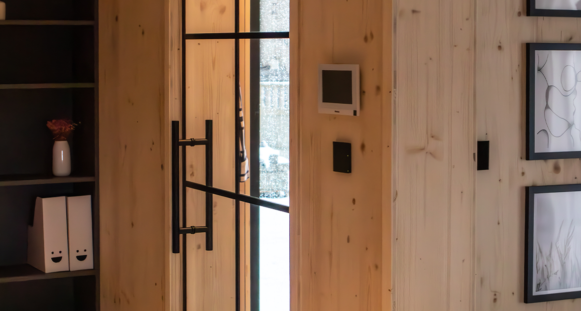 Eine Eingangstür aus Glas mit schwarzen Leisten und Griff. Draußen kann man sehen, dass es schneit. Die Innenwände sind komplett aus CLT- Holz in hoher Qualität. An der Wand neben der Tür ist ein Smart Home Display zu sehen.